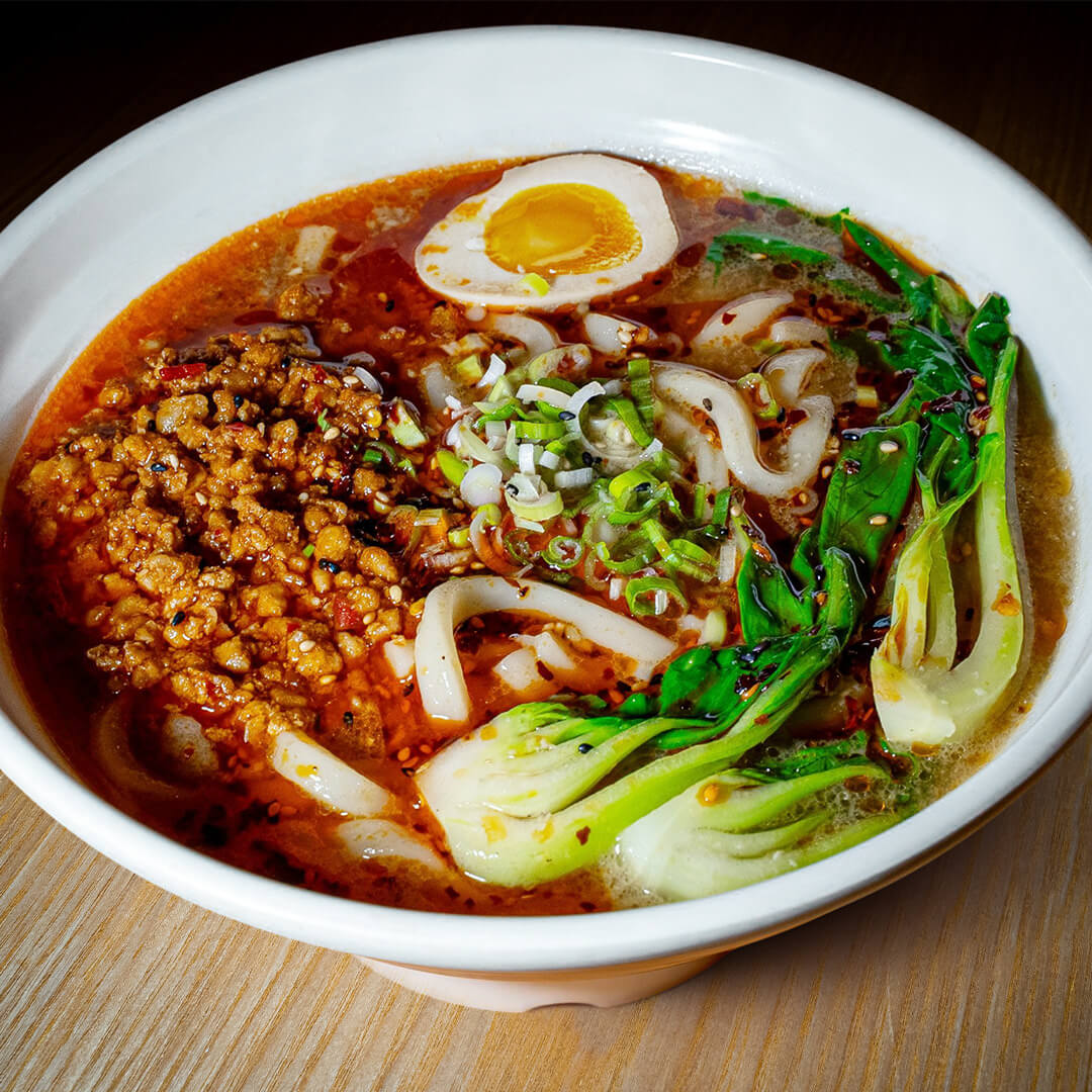 Dan Dan Udon Miami A vibrant bowl of spicy Dan Dan Udon from 107 Taste, showing ground pork, bok choy, a soft-boiled egg, and chili oil broth.